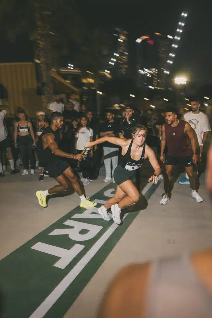 A relay race where a female runner in athletic clothing receives a baton from a male runner on a marked track at night, surrounded by other participants and spectators.