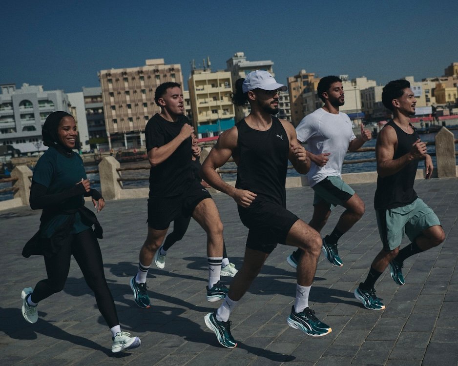 PUMA Running Dubai: A group of five people jogging on a paved walkway by the water, with buildings visible in the background. They are wearing athletic clothing and appear to be engaged in a group run or exercise routine.