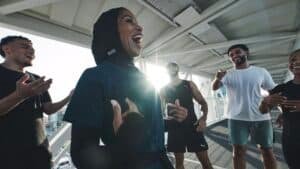 A group of people in athletic wear are laughing and engaging energetically with each other on a covered outdoor walkway in the sunlight.