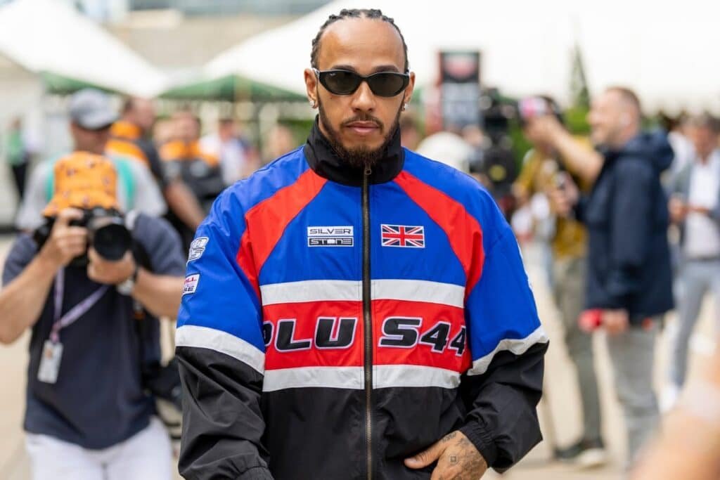 A man in sunglasses wearing a blue, red, and black jacket with British flag and "PLU S44" written on it, walks with people in the background.