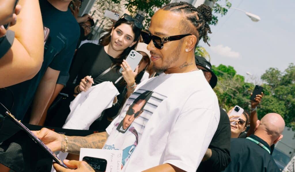 Lewis Hamilton in sunglasses and a printed Lewis Hamilton Plus44t-shirt signs an autograph amidst a crowd of people, some of whom are taking photos with their phones, outside on a sunny day.