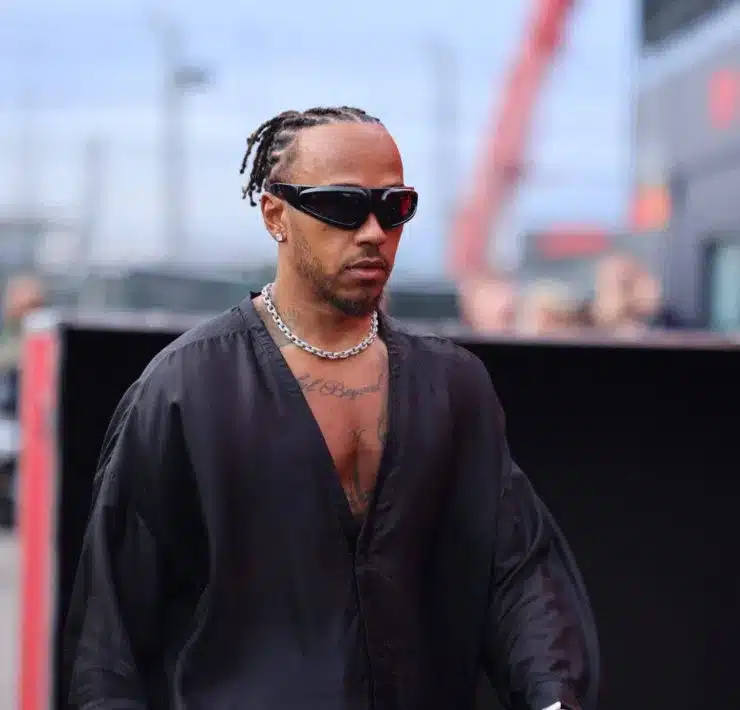 A person wearing sunglasses, a black shirt, and a silver chain is walking outdoors with braided hair and facial tattoos visible.