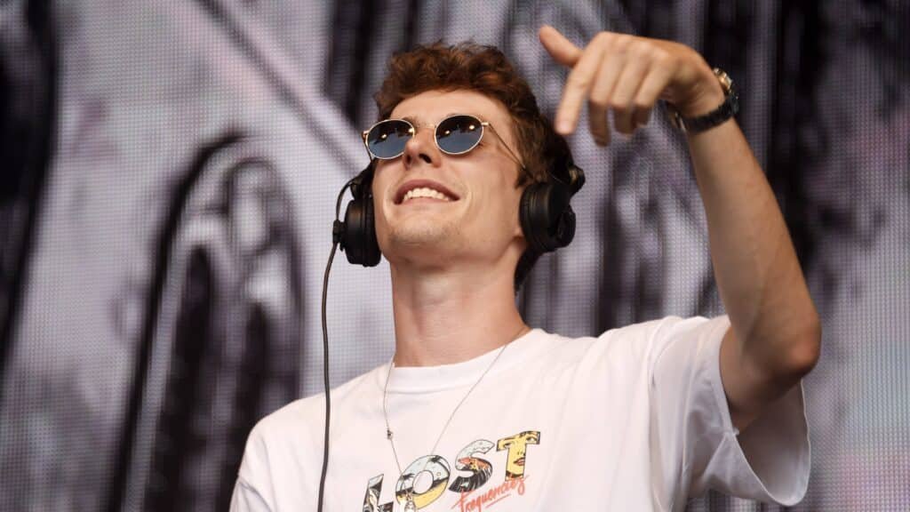 Lost Frequencies wearing sunglasses and headphones smiles while gesturing with their hand on stage, wearing a white T-shirt with 'Lost Frequencies' printed on it.