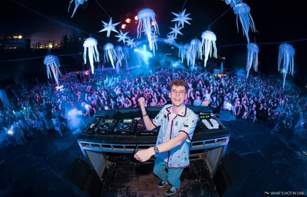 Lost Frequencies is energetically performing on stage at a nighttime outdoor music festival, surrounded by an enthusiastic crowd. The stage is adorned with inflatable jellyfish and star decorations hanging above, illuminated by colorful lights and effects.
