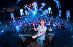 Lost Frequencies is energetically performing on stage at a nighttime outdoor music festival, surrounded by an enthusiastic crowd. The stage is adorned with inflatable jellyfish and star decorations hanging above, illuminated by colorful lights and effects.