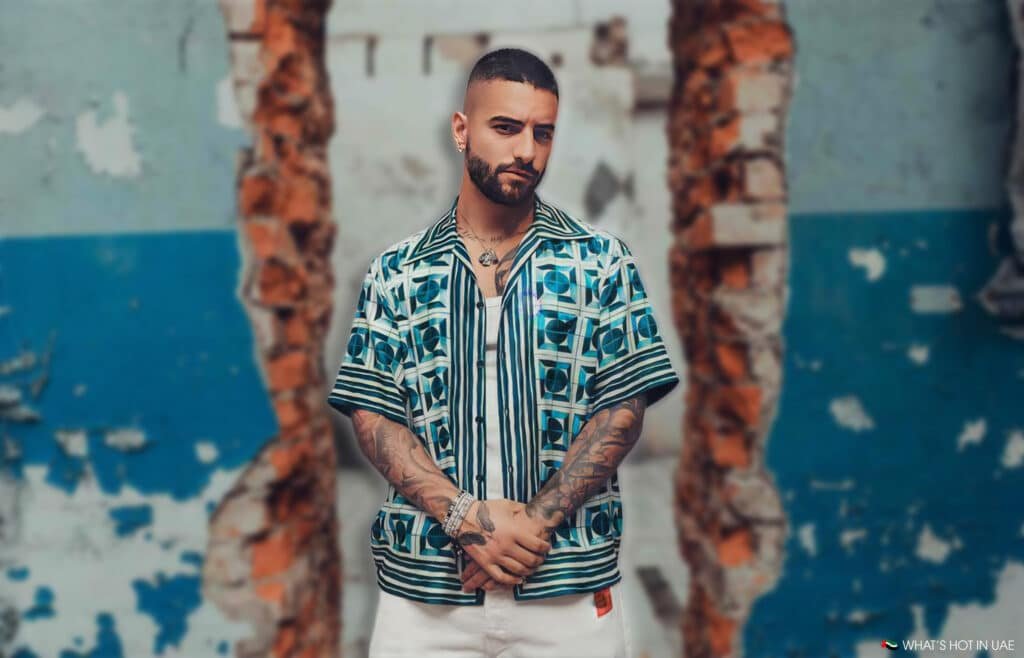 Maluma with tattoos on his arms stands in front of a weathered wall with peeling paint and exposed bricks. He is wearing a patterned short-sleeve shirt and white pants.
