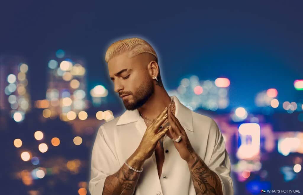 A person with platinum blonde hair and tattoos on their arms, wearing a white shirt and jewelry, stands against a blurred cityscape background at night.