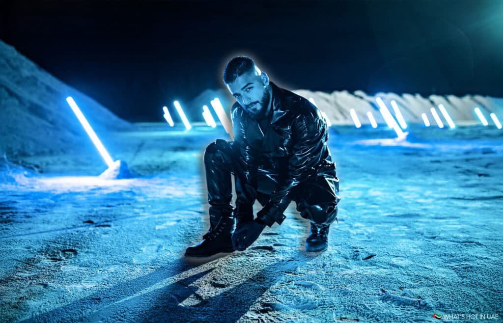 Maluma dressed in shiny, black clothing kneels on a futuristic landscape illuminated by neon blue lights, surrounded by dunes and bright light rods sticking out of the ground.