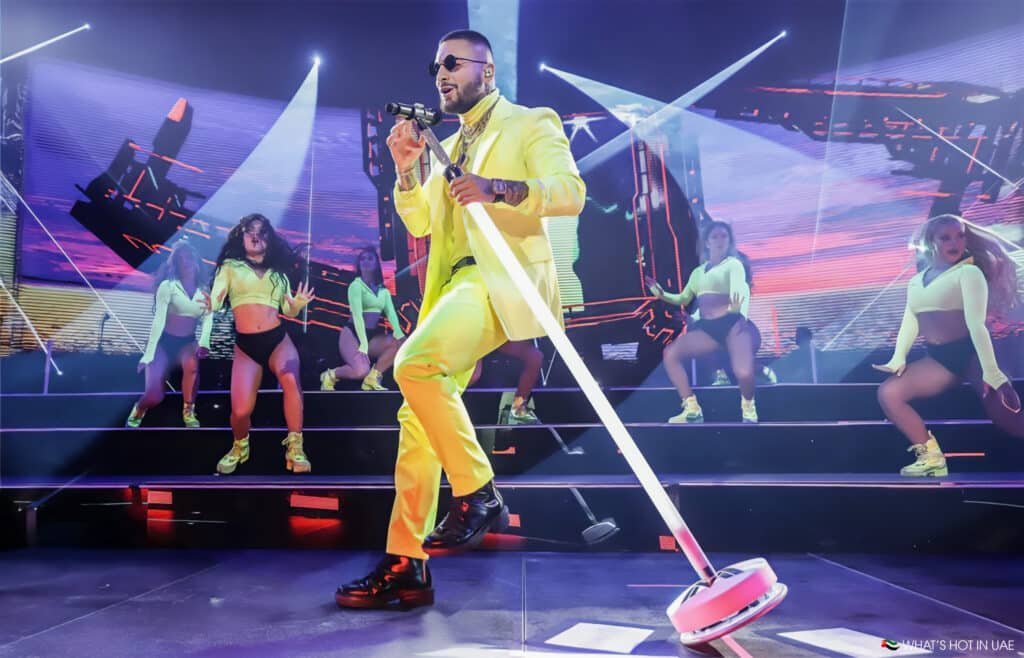 A performer wearing a bright yellow suit sings on stage with a microphone, surrounded by four dancers in neon yellow outfits and black shorts. The stage is lit with vibrant lights and a digital backdrop, creating an energetic atmosphere.