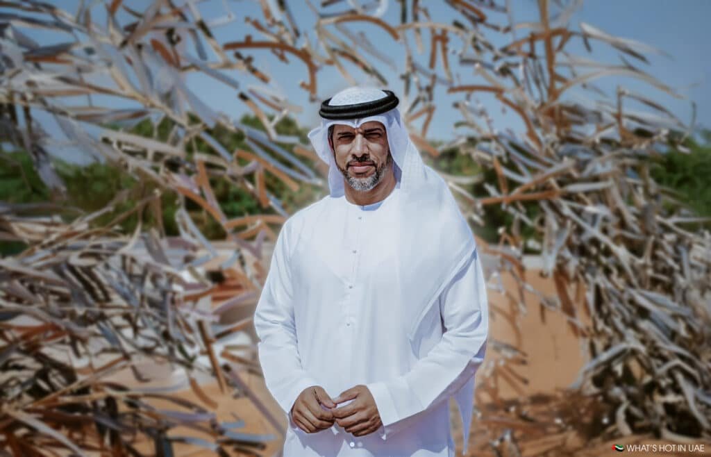 A person in traditional Emirati attire stands outdoors with a background of abstract sculptural elements and greenery.