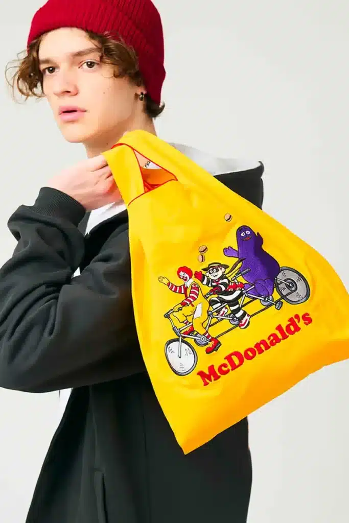 A person wearing a red beanie and a black jacket holds a bright yellow bag with McDonald's branding and cartoon characters riding a tandem bike printed on it.
