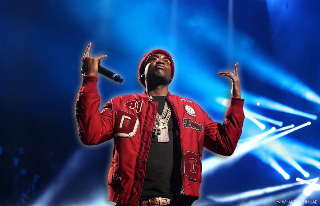 Meek Mill in Dubai wearing a red jacket and beanie, performing on stage with dramatic lighting effects in the background.