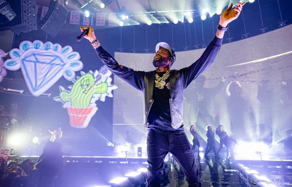 A performer on stage with arms raised, holding a microphone, surrounded by bright lights and cartoonish inflatable figures, including a diamond and a cactus.