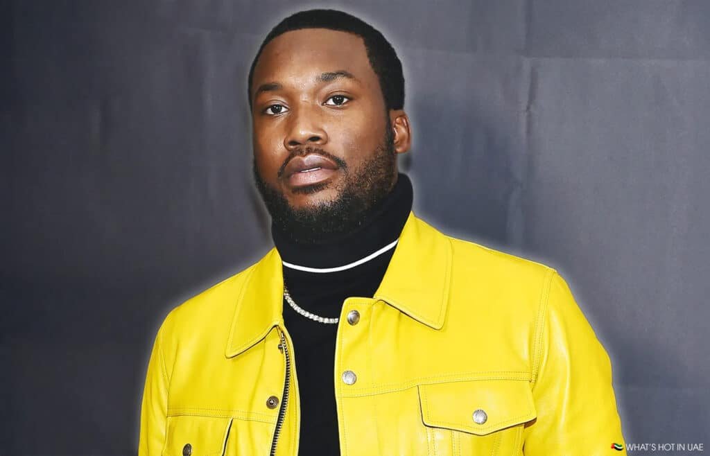 Meek Mill in Dubai wearing a bright yellow jacket over a black turtleneck with a necklace, standing against a dark background.