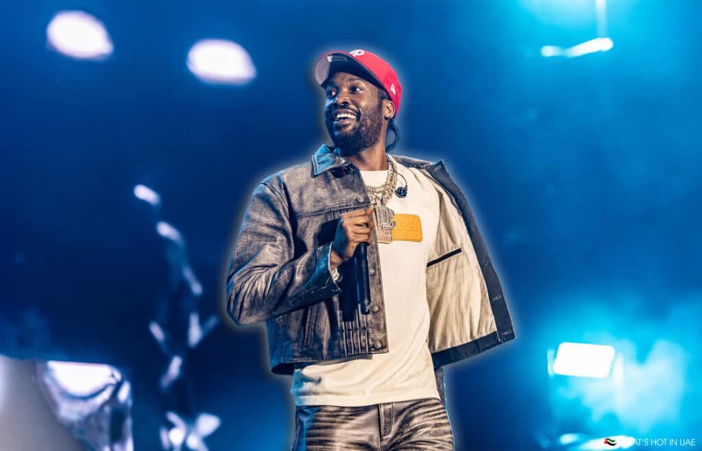 A performer on stage wearing a denim jacket, white T-shirt, and red cap, holding a microphone with bright stage lights in the background.