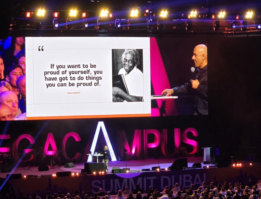 Megacampus Summit Dubai 2026: A large stage with a big screen displaying a quote by Oseola McCarty, accompanied by her photo. The quote reads, 'If you want to be proud of yourself, you have got to do things you can be proud of.' A speaker stands to the right of the screen addressing the audience, with the words 'MEGACAMPUS SUMMIT DUBAI' visible in the background.
