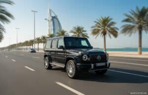 A black Mercedes-Benz G-Class SUV driving on a wide road lined with palm trees, with the Burj Al Arab in the background against a clear blue sky in Dubai, UAE.