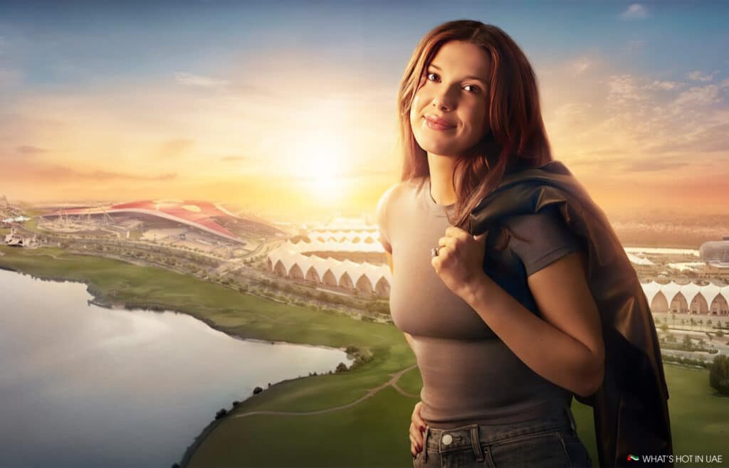 A woman stands smiling with a sunset-lit landscape in the background, featuring a large, modern red-roofed building near a body of water and green fields.