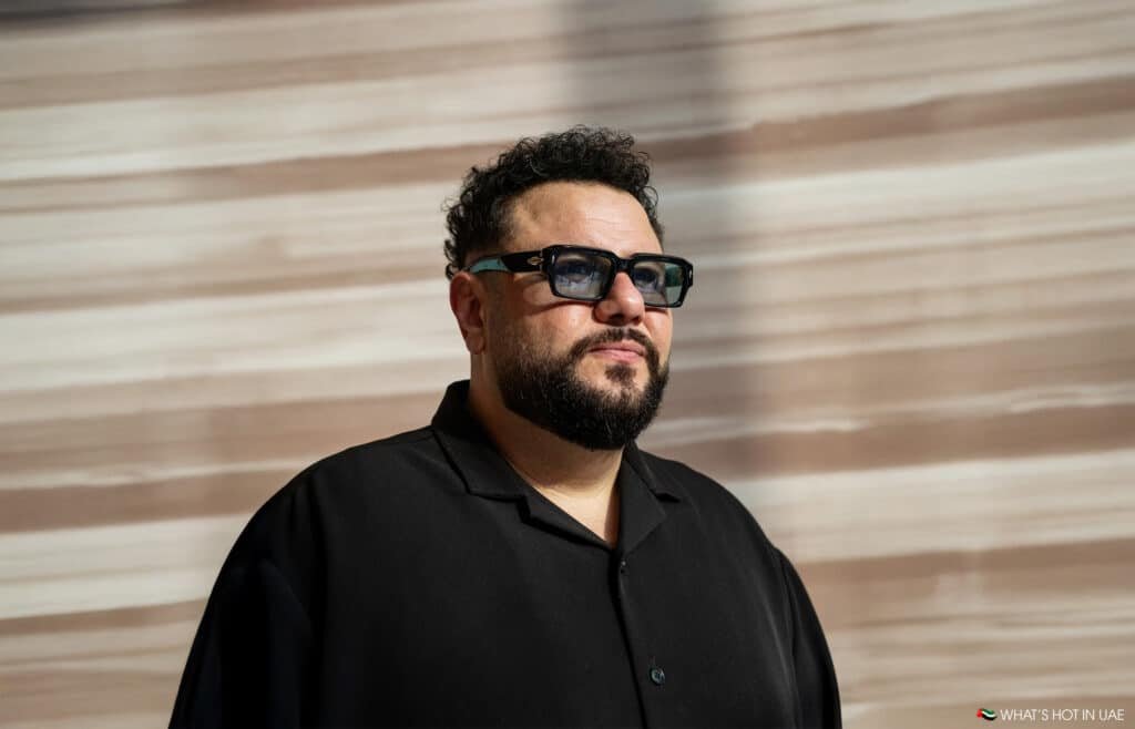 Mo Amer wearing dark sunglasses and a black shirt stands against a blurred background with horizontal stripes.