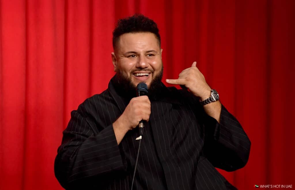 A man with a beard smiling and holding a microphone while gesturing with his hand, as if talking on the phone, against a red curtain background.