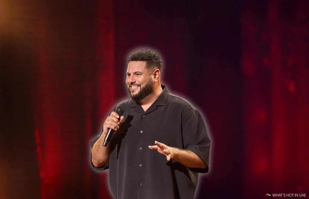 A man holding a microphone is performing on stage with a red curtain backdrop, wearing a black button-up shirt and smiling.