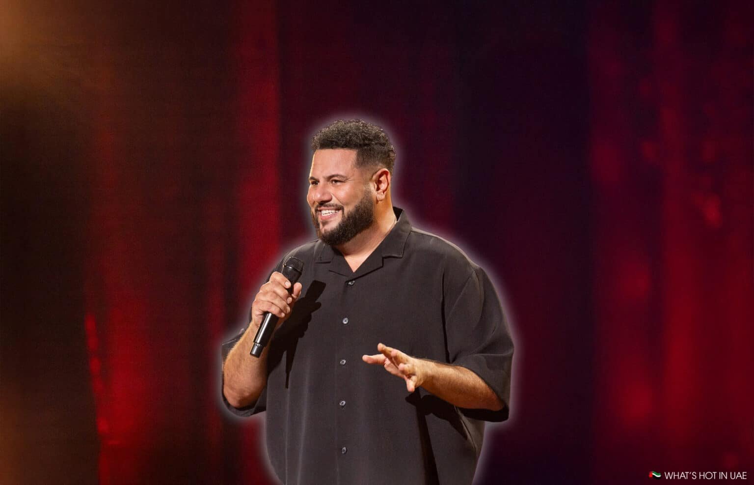 A man holding a microphone is performing on stage with a red curtain backdrop, wearing a black button-up shirt and smiling.