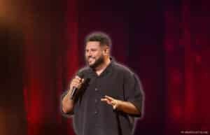 A man holding a microphone is performing on stage with a red curtain backdrop, wearing a black button-up shirt and smiling.