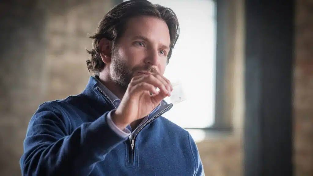 Bradley Cooper with a beard, wearing a blue zippered sweater, holds a small transparent bag of NZT-48 with his right hand. He is looking intently at the bag in a softly lit indoor setting with a blurred background.