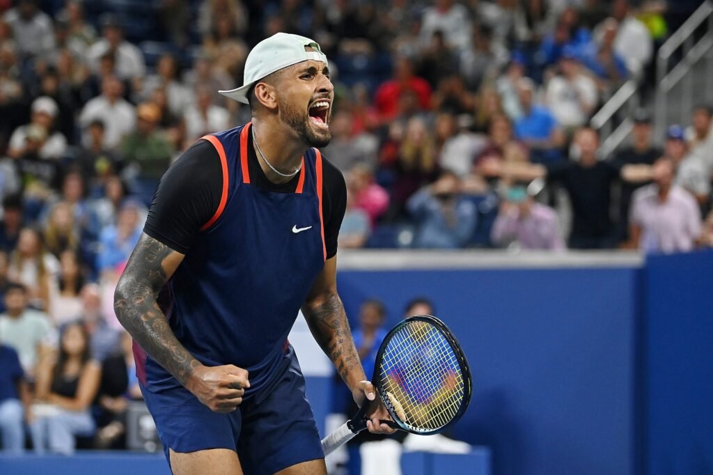 A tennis player wearing a blue and black outfit and a backwards cap is celebrating passionately on a court, holding a tennis racket, with a cheering crowd in the background.