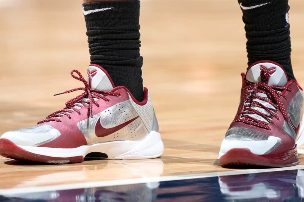 Close-up of a pair of Nike Kobe 5 Protro 'Lower Merion Aces' basketball shoes with maroon and silver colors on a basketball court. The shoes feature a swoosh logo and are worn with black socks.