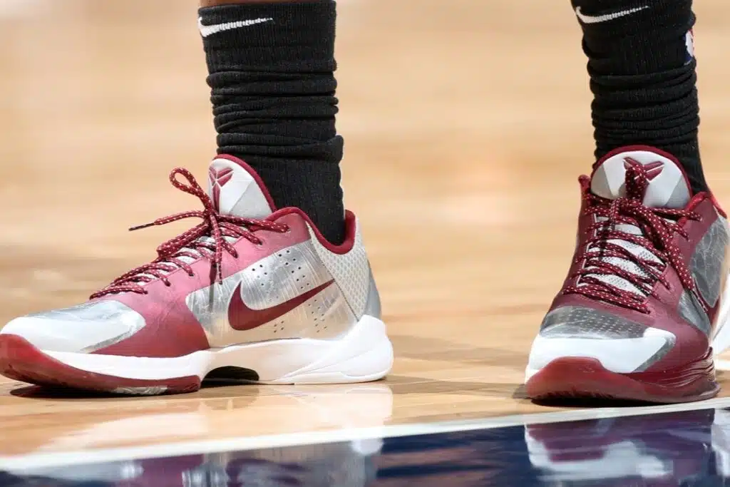 Close-up of a pair of Nike Kobe 5 Protro 'Lower Merion Aces' basketball shoes with maroon and silver colors on a basketball court. The shoes feature a swoosh logo and are worn with black socks.