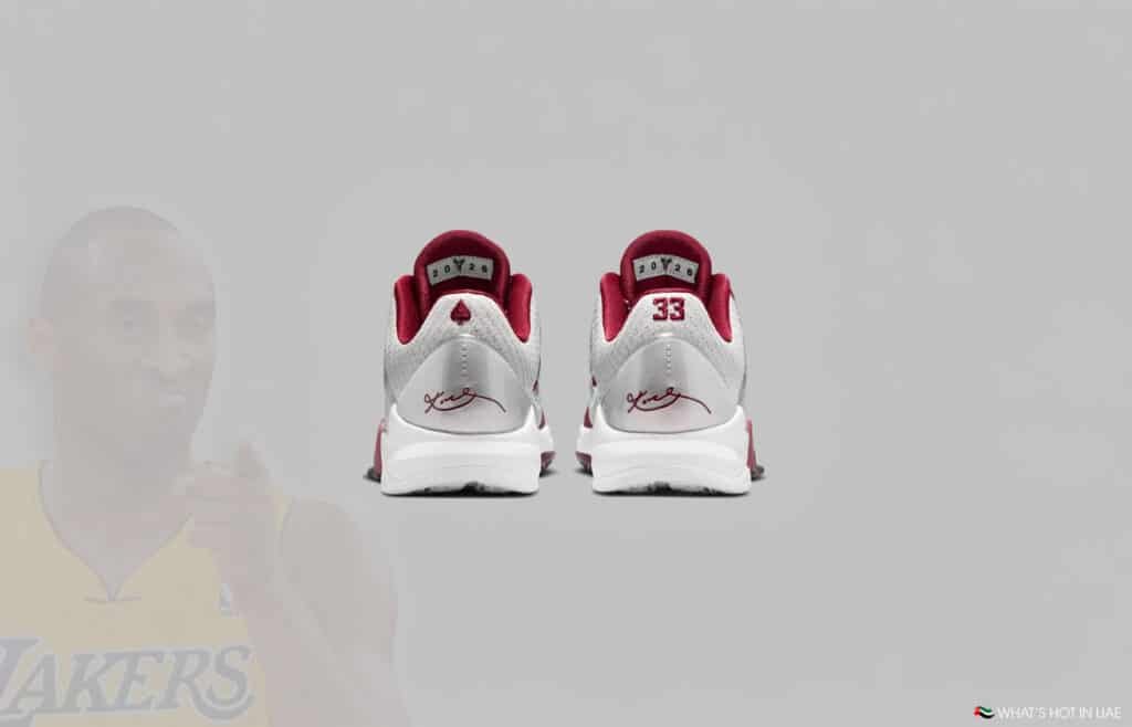 Back view of a pair of sneakers with white and burgundy colors, featuring a signature and a number 33 on the heels. A faded image of a basketball player in a Lakers jersey is in the background.