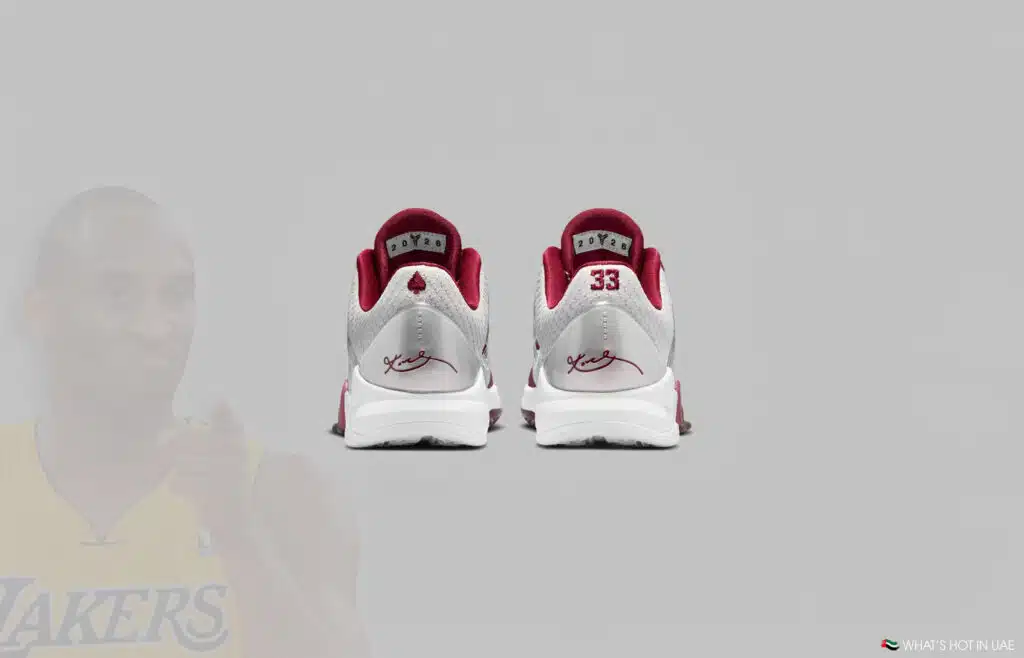 Back view of a pair of sneakers with white and burgundy colors, featuring a signature and a number 33 on the heels. A faded image of a basketball player in a Lakers jersey is in the background.