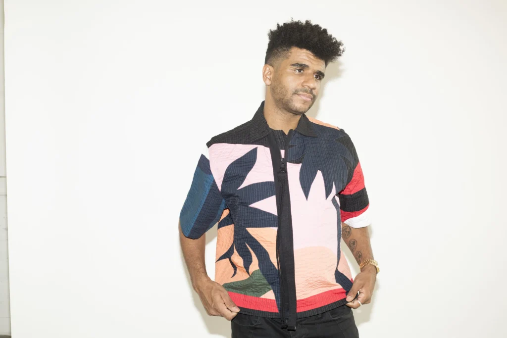 Jamie Jones at Be Beach Dubai standing against a plain white background, wearing a colorful, abstract-patterned shirt with shades of black, pink, red, and blue, and black pants. The person has curly hair and visible tattoos on their left arm.