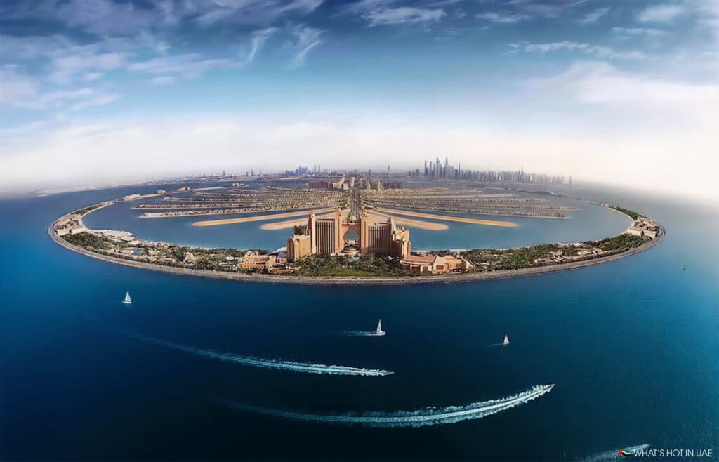 Aerial view of the Palm Jumeirah in Dubai, showcasing the iconic palm-shaped artificial archipelago featuring the Atlantis Hotel at the tip, surrounded by blue ocean waters and the city skyline in the background.
