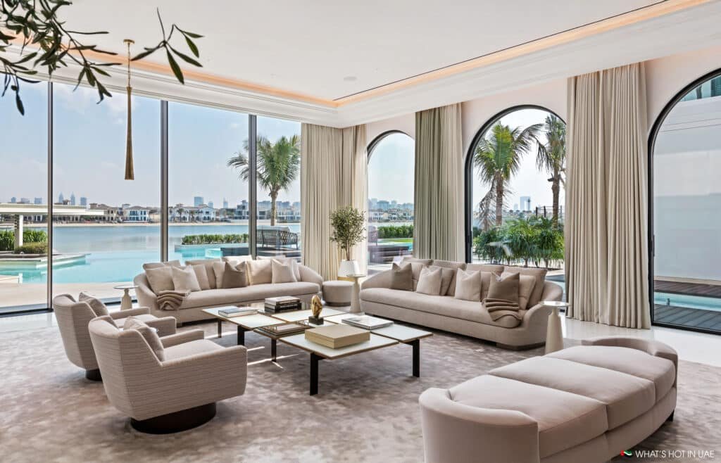 Luxurious living room with large sofas and armchairs arranged around a central coffee table, featuring floor-to-ceiling windows offering a view of a pool and palm trees.