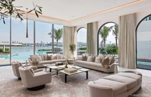 Luxurious living room with large sofas and armchairs arranged around a central coffee table, featuring floor-to-ceiling windows offering a view of a pool and palm trees.