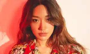 A woman with long hair wearing a colorful patterned shirt, illuminated by warm red lighting, looking directly at the camera.