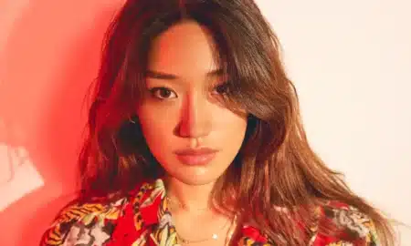 A woman with long hair wearing a colorful patterned shirt, illuminated by warm red lighting, looking directly at the camera.