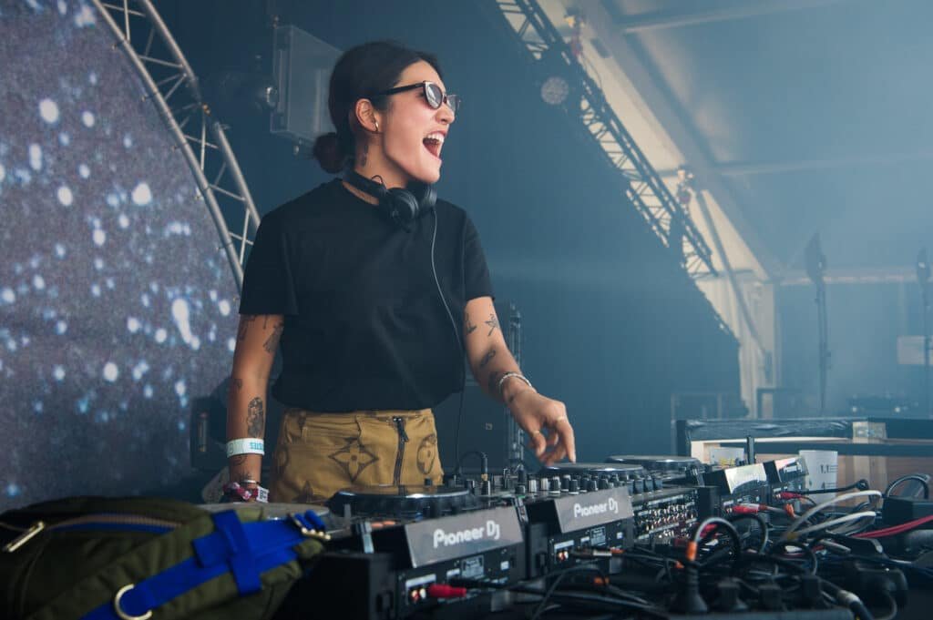 Peggy Gou DJ with headphones around their neck, wearing sunglasses and casually dressed, enthusiastically performing at a music festival with DJ equipment in front of them and a large screen displaying a starry visual.