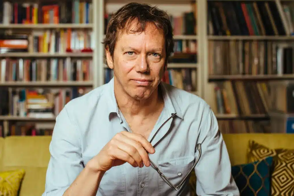 Robert Greene in a light-colored shirt holding glasses, sitting in front of packed bookshelves.