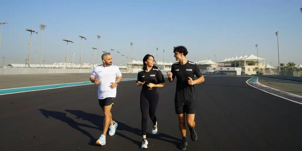 Three people jogging on Yas Island race track, with two men wearing sportswear and a woman in the middle, under a clear blue sky.