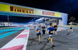 A group of people jogging on a racetrack at night under a Pirelli banner, with modern buildings and palm trees in the background.