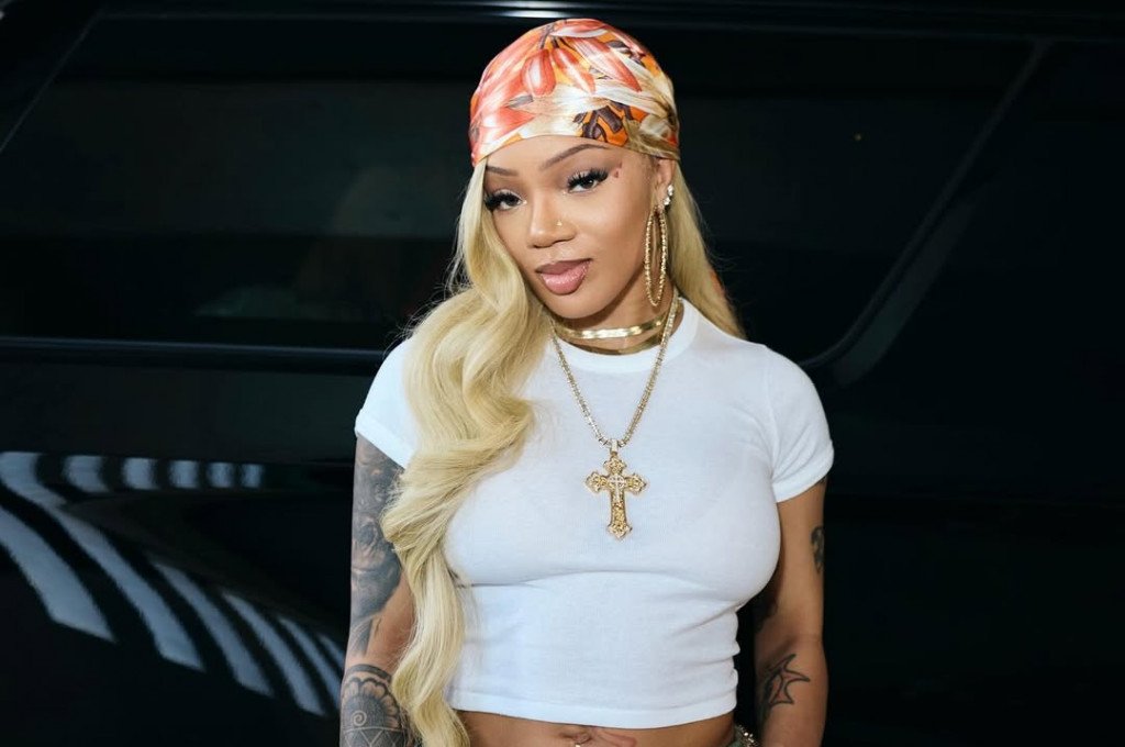 A woman with long, wavy blonde hair wearing a colorful headscarf, a white crop top, gold hoop earrings, and a gold cross necklace, standing in front of a dark vehicle.