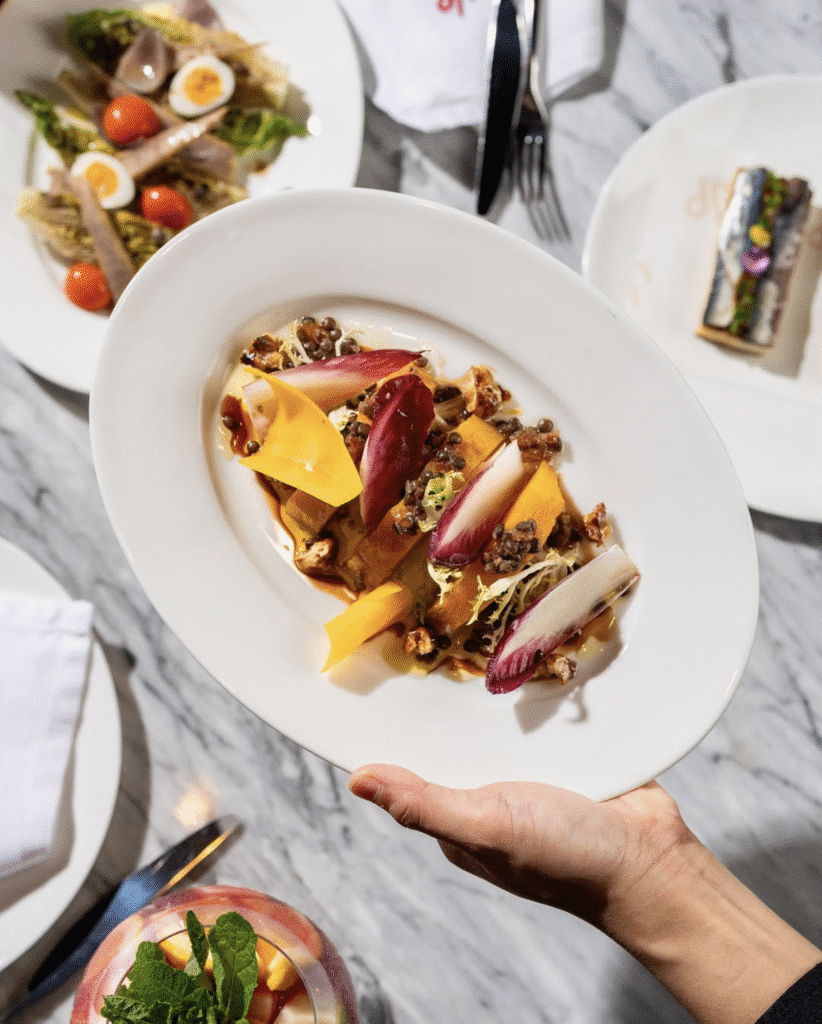 The 8 Best Restaurants in Abu Dhabi: A hand holding a plate of colorful gourmet salad featuring endive leaves, cheese slices, and nuts, with other dishes and a drink in the background on a marble surface.