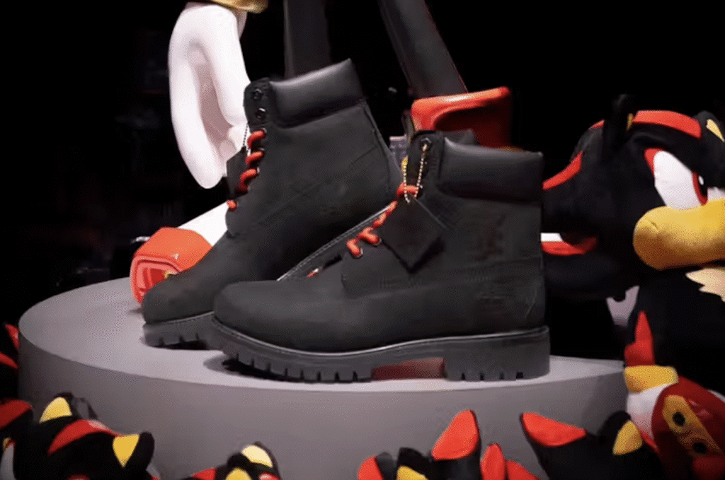 A pair of black Timberland Shadow the Hedgehog Boots with red laces and a tag displayed on a round platform, surrounded by plush toys featuring black and red details.