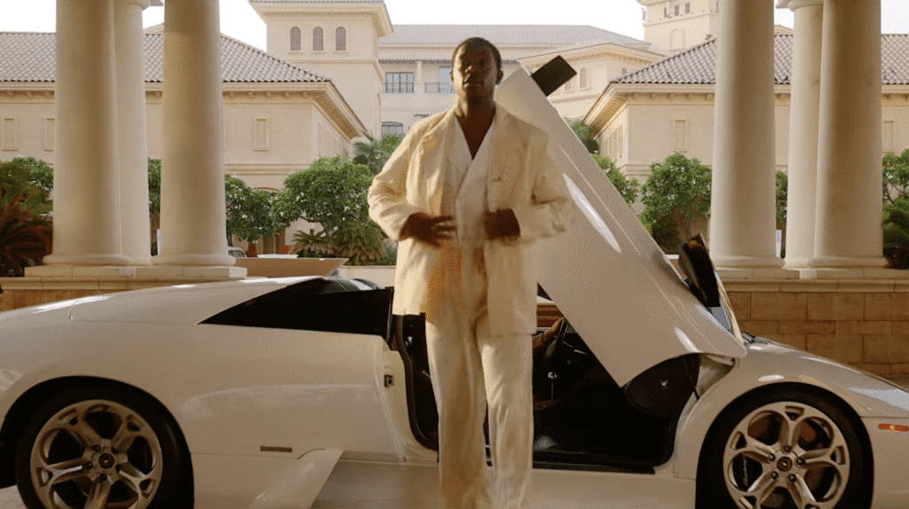 A person dressed in a white suit stands beside an open door of a white luxury car in front of a grand building with columns and lush greenery.
