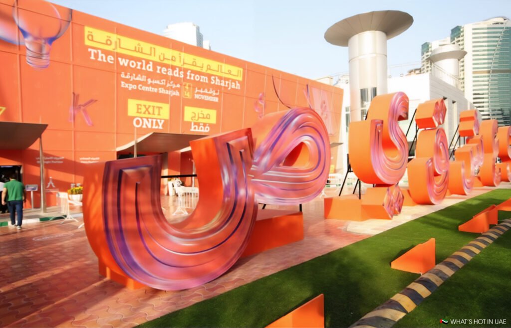 A large, colorful 3D lettering installation in Arabic is displayed outside the Expo Centre Sharjah. The building features orange walls with signage advertising a reading event called 'The world reads from Sharjah'. People are entering the venue through a nearby entrance.
