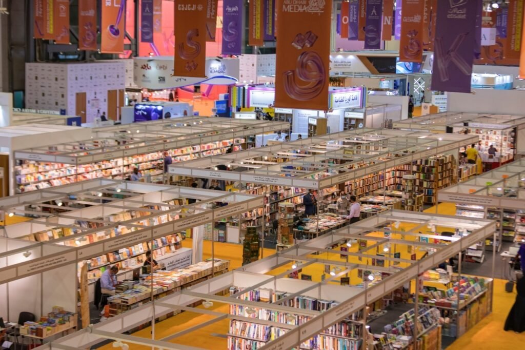 A large indoor book fair with numerous stalls displaying books, people browsing, and colorful banners hanging from the ceiling.