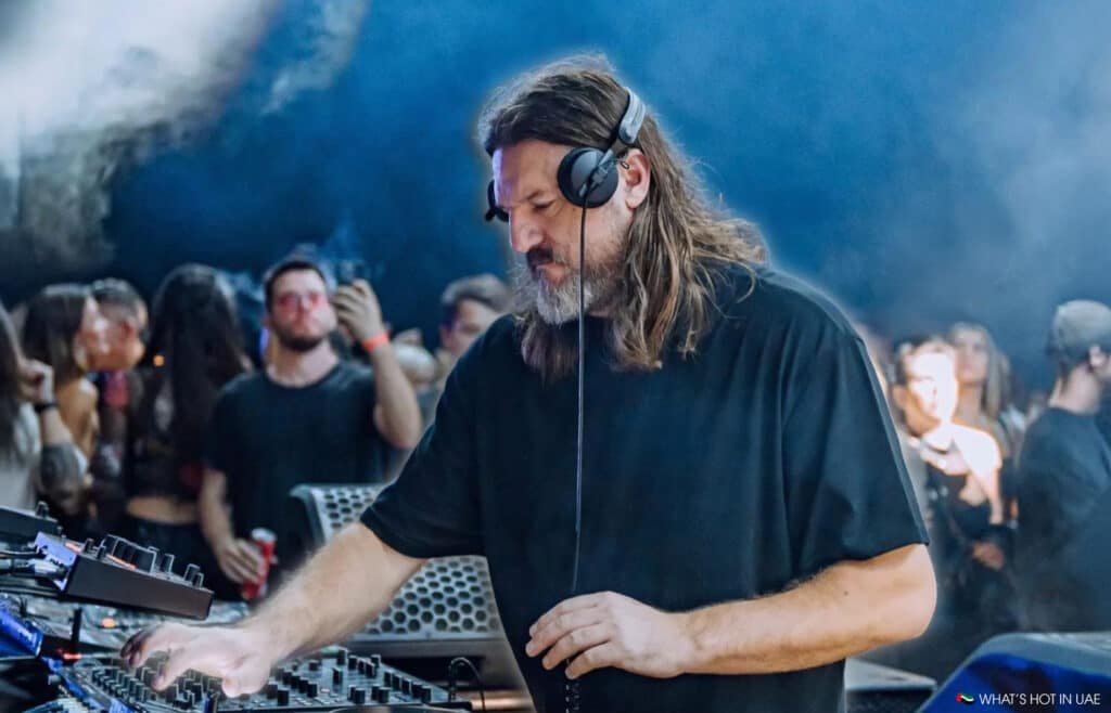 A DJ with long hair and a beard, wearing headphones, is intensely focused on mixing music at an electronic music event with a crowd in the background.